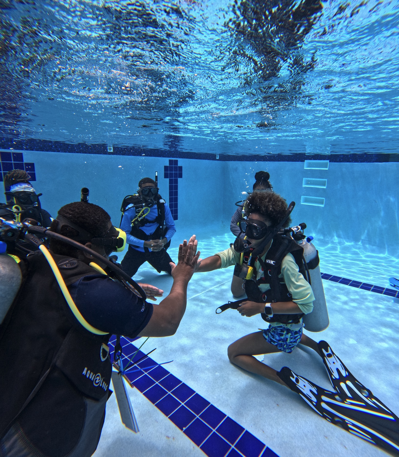 IDC students practice confined water skills underwater in a pool, with two divers giving each other a high-five while instructors observe